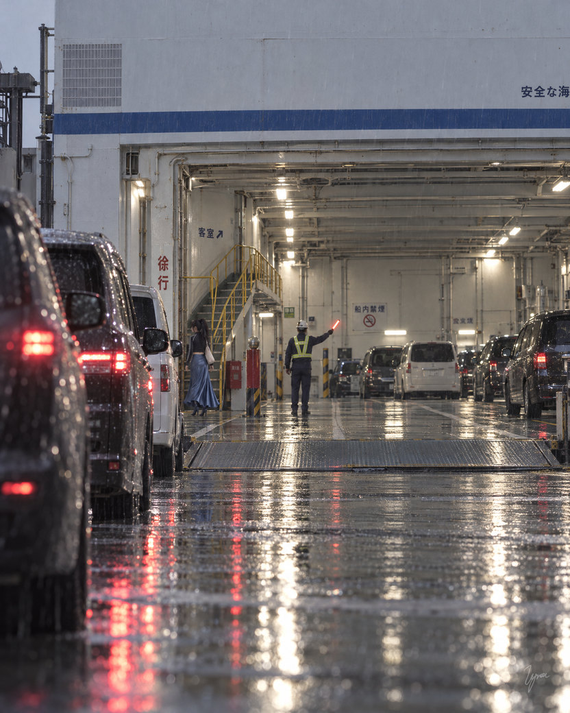 From a rainy quay at dusk, past a line of cars with red tail lights waiting to board. The ferry’s open stern shows the lit car deck; deep inside, the deck hand signals with his baton and the young woman in the blue skirt is a small figure near the stairs.
